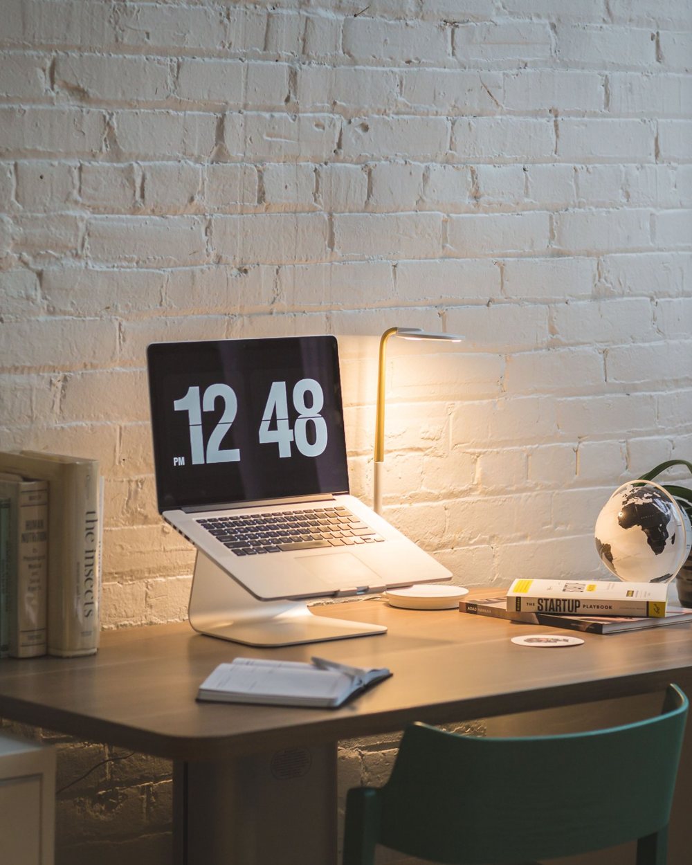 A warmly lit desk with a laptop on a stand, a lamp and a plant